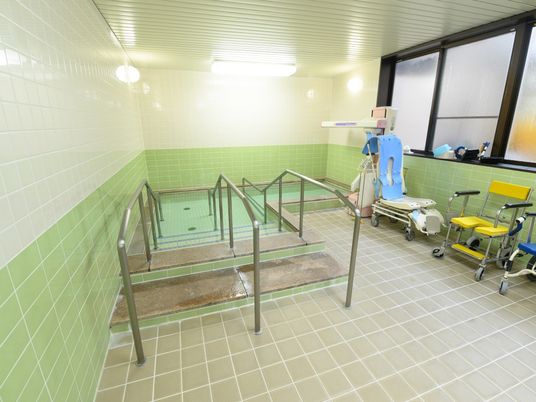 施設の写真 浴槽の前に、２名ずつ利用できる階段があり、両手でつかまれる手すりがある。リールのついた椅子がいくつか置かれている。