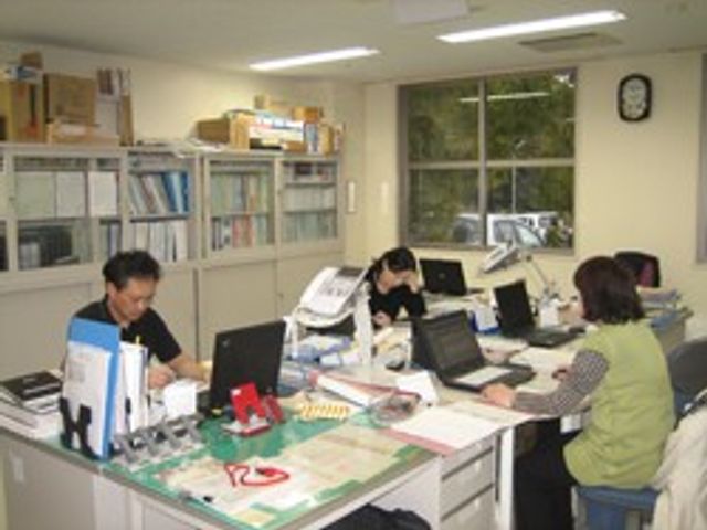 スタッフが働く事務室 スタッフイメージ 広々としたデスクでチームが仕事に集中しています。自然光が差し込む、落ち着いた空間です。