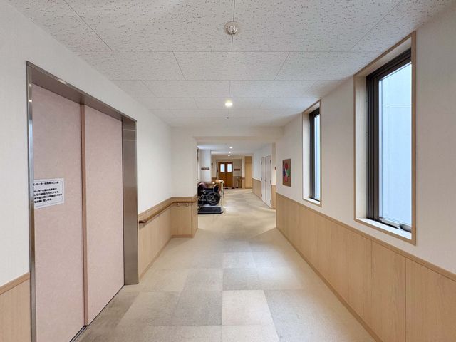 明るい廊下とエレベーター前 廊下   手すりが続く明るい廊下で、窓から自然光も入っています。奥に車椅子が見え、移動しやすさが伝わります。