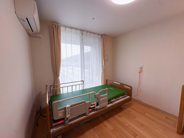 明るい居室のベッド 居室   大きな窓から自然光が入り、木目調の床が落ち着いた印象です。電動ベッドが置かれ、介助しやすい空間に見えます。