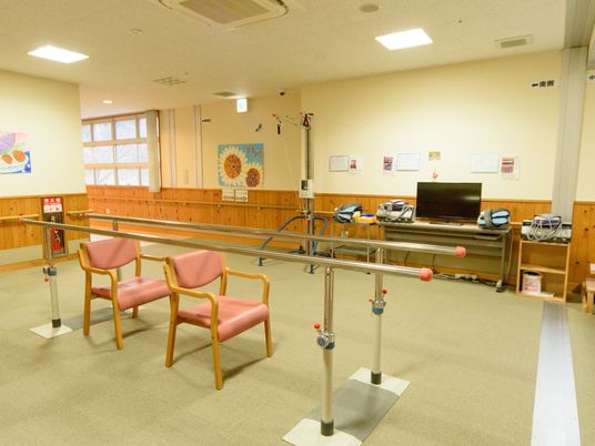 通路途中に設けられている機能訓練室で、平行棒の間に椅子が２脚置かれている。壁には、手すりが取り付けられている。