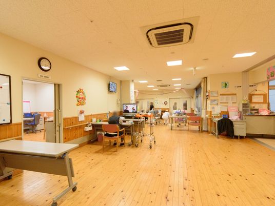 テレビが設置されている広々とした廊下で、入居者様が椅子に座ってくつろいでいる。奥には、リビングスペースが見える。