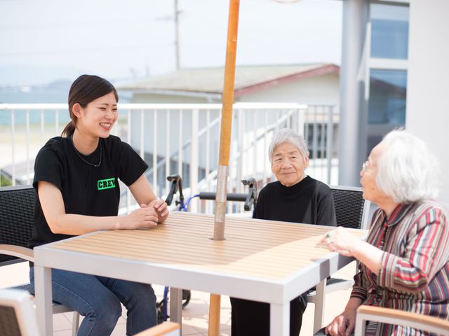 屋外テーブルで会話する人々 笑顔でコミュニケーションを取るスタッフと利用者様の姿。明るい天気の下で楽しむひとときが垣間見えます。