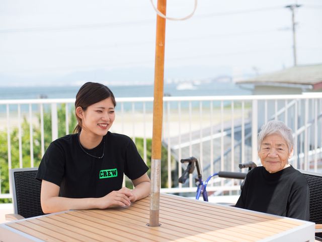 テラスで会話するスタッフと利用者 潮風を感じる開放的なテラスで、利用者とスタッフが心地よい時間を過ごしています。スタッフの明るい表情が印象的です。