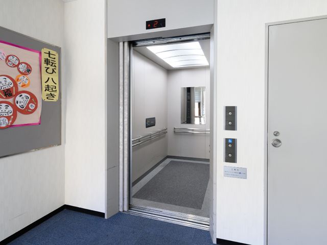 廊下のエレベーター口 エレベーター ゆとりあるスペースのエレベーターホールは、どなたでも快適に利用できるようになっています。明るい照明と落ち着いた色合いの壁が印象的です。