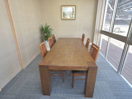 施設の写真 大きな窓があり、床にじゅうたんが敷かれている部屋である。木のテーブルがあり、3人ずつ向かい合って座ることができる。壁には絵が飾られている。