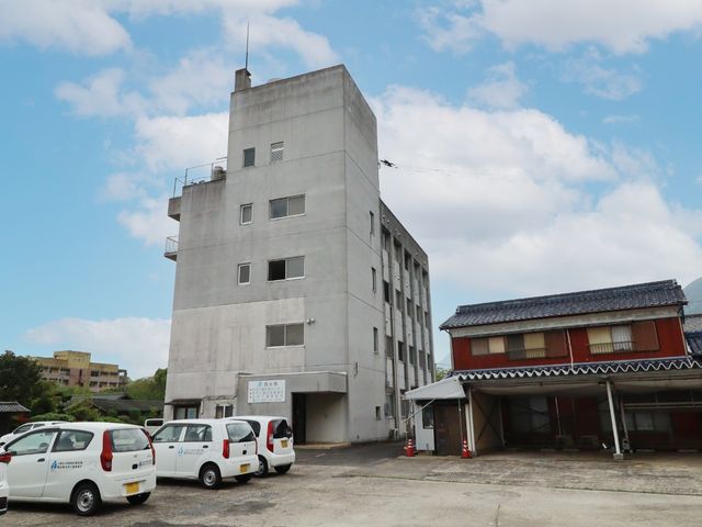 施設の外観 外観   青空の下に建つ多層の建物で、駐車スペースには送迎車が並んでいます。木造の建物も隣接しており、外回りの様子が分かります。