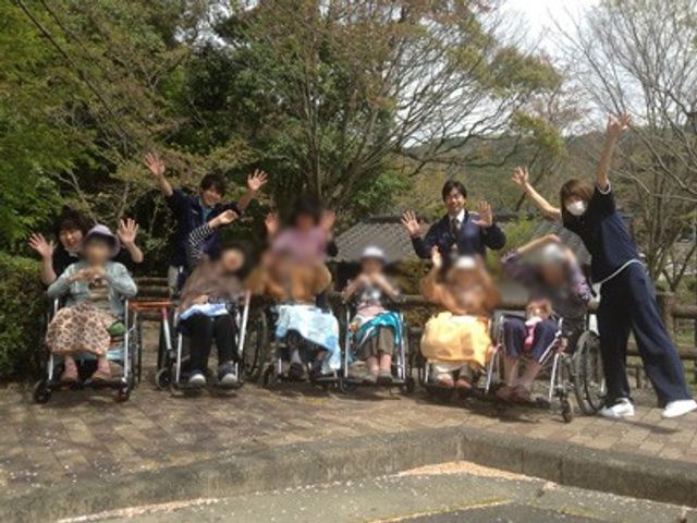 元気な人々が集う場所 庭 笑顔で手を振る利用者とスタッフが、自然豊かな庭で和やかに過ごしています。