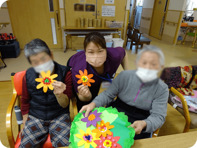 工作活動を楽しむ様子 多目的室 入居者様がスタッフと一緒に手作りの花飾りを楽しんでいます。楽しさが伝わる活動の様子です。