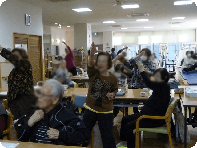 体操をする皆さんの様子 機能訓練室 広々とした空間で、利用者がリラックスして体操を楽しんでいます。温かな雰囲気が漂っています。