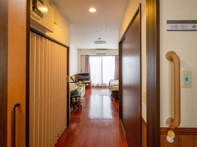 廊下から見る居室入口 サポート 私たちの働く場所は、居心地の良さを大切にしている居室があります。温かみのある木目調に、ゆったりと過ごせる広さです。