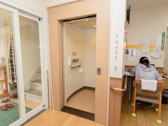 居室の入口と利用者 エレベーター ゆとりある居室入口から、一人の利用者がくつろぎの時間を過ごしている様子が見て取れます。安全に配慮された設計で、快適な日々を送れる環境が整っています。