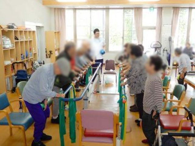 体操をする人々の部屋 機能訓練室 明るい部屋でリハビリ体操を行っており、窓から自然光が差し込みます。スタッフが丁寧にサポートしています。