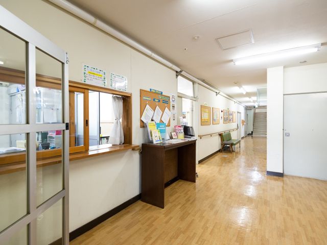 廊下と受付の空間 エントランス ゆったりとした通路に面した受付は、来訪者の対応をスムーズに行えるよう配慮されています。周囲は掲示物で情報が豊富にあり、明るく開放的な雰囲気を感じることができます。