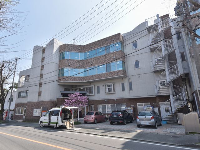 快適そうな建物の外観 外観 ゆとりを感じる建物前には、明るい日差しの下で入居者の対応にあたるスタッフの姿が見られます。複数の乗用車が停まっており、来訪者にも配慮した設計となっています。