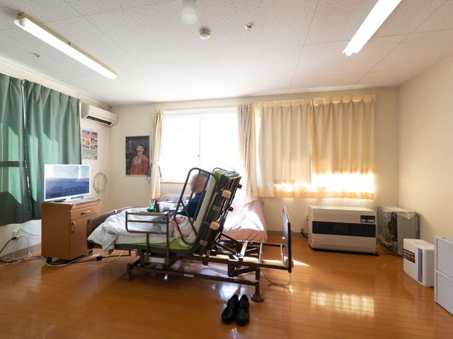 居室にあるベッドとテレビ 居室 ゆったりとした居室にはリクライニングベッドが完備され、快適に過ごせるよう配慮されています。窓からは自然光が差し込み、温かみのある雰囲気を提供しています。