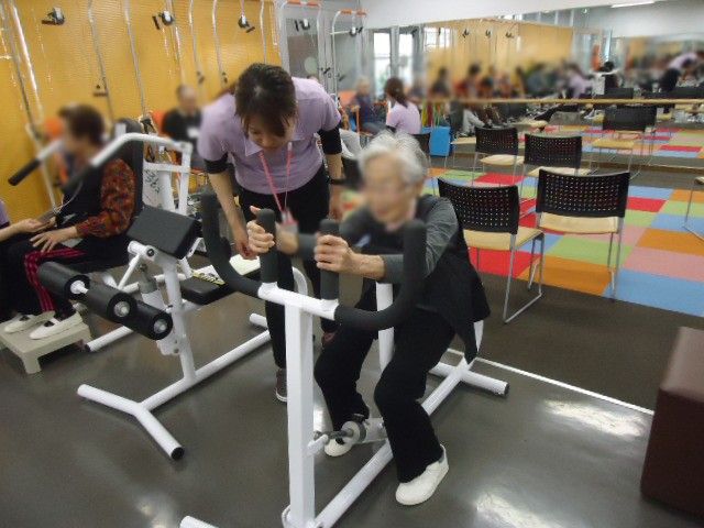 トレーニング指導中の様子 機能訓練室 スタッフが利用者に優しく指導しています。色とりどりの床が特徴的な訓練スペースです。