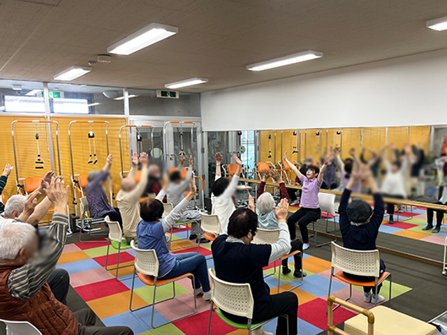 体操をする人々の姿の機能訓練室 機能訓練室 明るい空間の中、利用者が元気に体操を楽しんでいます。和やかな雰囲気の中で指導が行われています。