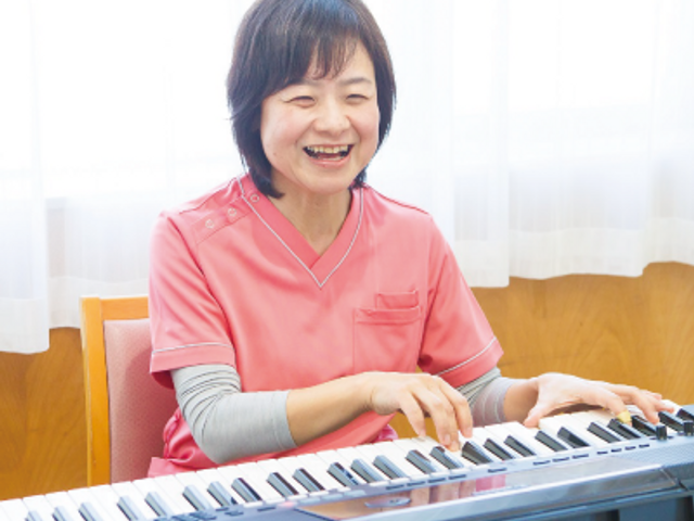 笑顔でピアノを弾くスタッフ リビング 明るい笑顔でピアノを奏でるスタッフがいる居心地の良いリビングです。音楽を通じて日々の潤いを提供しています。