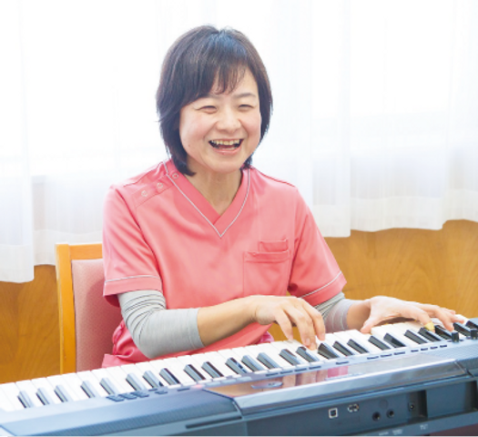 笑顔でピアノを弾くスタッフ リビング 明るい笑顔でピアノを奏でるスタッフがいる居心地の良いリビングです。音楽を通じて日々の潤いを提供しています。