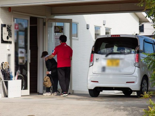 車到着時の出入口 エントランス 車から降りた利用者を職員が温かく迎え入れる場面です。家庭的な雰囲気で安心感があります。