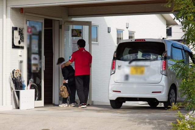 車到着時の出入口 エントランス 車から降りた利用者を職員が温かく迎え入れる場面です。家庭的な雰囲気で安心感があります。