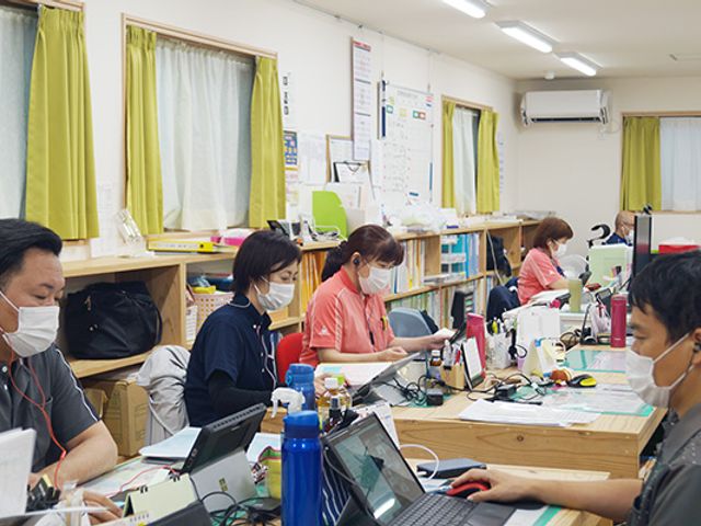スタッフが働く執務室 スタッフイメージ マスクを着けた職員の方々が、それぞれのデスクで忙しく作業をされています。連携を取りながら、日々の業務に励んでいる様子が伺えます。