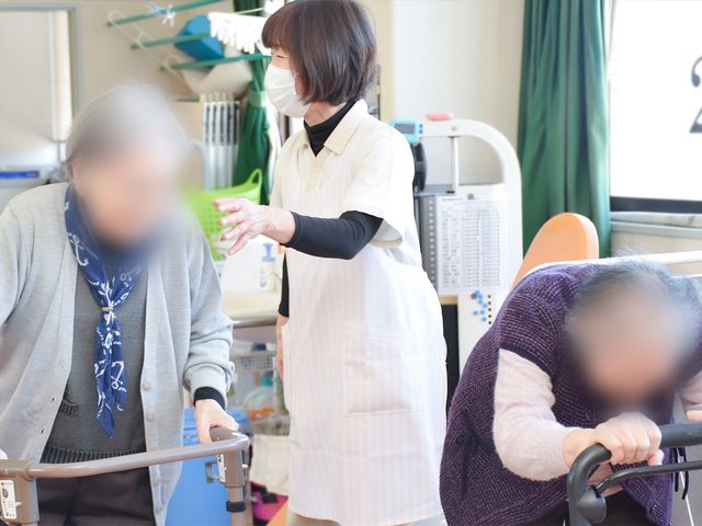 介助風景の室内 機能訓練室 自然光が差し込む中、介護士が高齢者を優しくサポートしています。安心して働ける環境です。