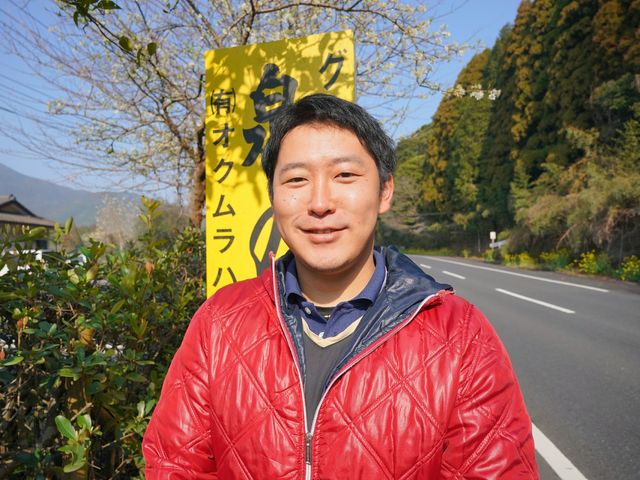 笑顔の人物と黄色い看板 スタッフ ニコリと笑顔を見せる方が、明るい黄色の看板の前に立っています。周囲は自然に囲まれ、居心地が良さそうな雰囲気です。