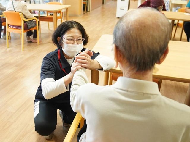 スタッフと利用者の手をつなぐ場面 居室 ゆるやかな時間が流れる居室で、スタッフが笑顔で利用者さまの手を握っています。安心できる雰囲気が伝わります。