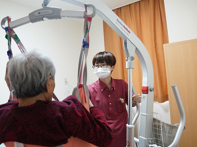 介護スタッフと利用者の機能訓練室 機能訓練室 笑顔で優しく支えるスタッフと、リハビリ機器を使用して体を動かす高齢の方がいらっしゃいます。安心して日々の体力維持に励める環境です。