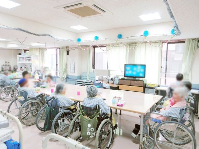 明るく広い空間の共有スペース リビング 自然光が差し込む広々としたリビングです。皆さんが車椅子で集まり、穏やかな時間を過ごしています。
