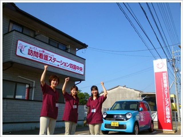 看板前で手を上げるスタッフと車 外観   看板の前にスタッフが3人並び、車両も見えます。就業時の雰囲気を想像しやすい一枚です。