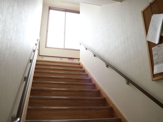 上の階へと続く階段がある。階段の右側には掲示板があり、紙が貼りつけられている。両側には手すりが設置されている。