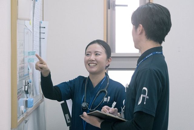 スタッフが掲示物を指さす様子 笑顔でコミュニケーションを取りながら、仕事の流れを確認する風景です。明るい雰囲気の中での協力が、日々の業務を支えています。