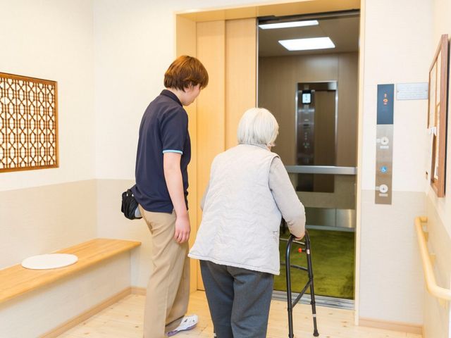 廊下に設置されたエレベーター 介護 ケアを提供するスタッフが、歩行補助器を使う利用者様と一緒にエレベーター前で待っています。安心して移動できる設計が考えられた廊下です。