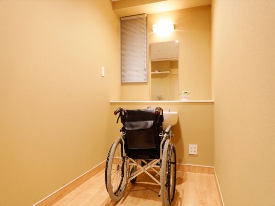 施設の写真 コの字型スペースの真ん中に、１台の車椅子が置いてある。その前方に、長方形の鏡と明るいライトが取りつけられている。