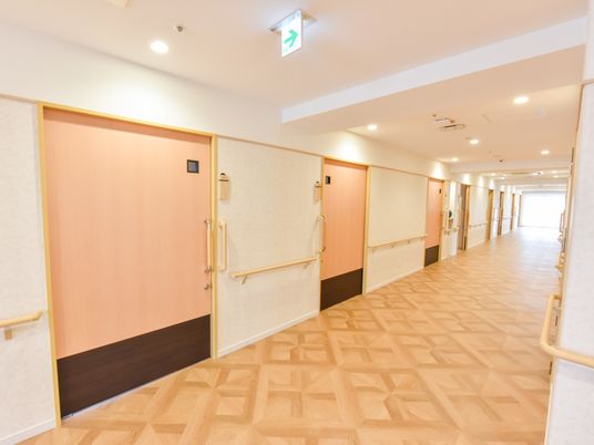 施設の写真 明るい木目調の床と生成色の壁紙に、やさしいピーチ色の扉が、あたたかみのある、上品で落ち着いたコーディネイトの空間である。