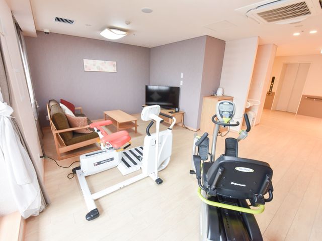 リハビリ器具とソファの居室 共有スペース 広々とした居室には、リハビリ用の器具が設置され、日常生活のサポートが整っています。ソファでのくつろぎも可能です。