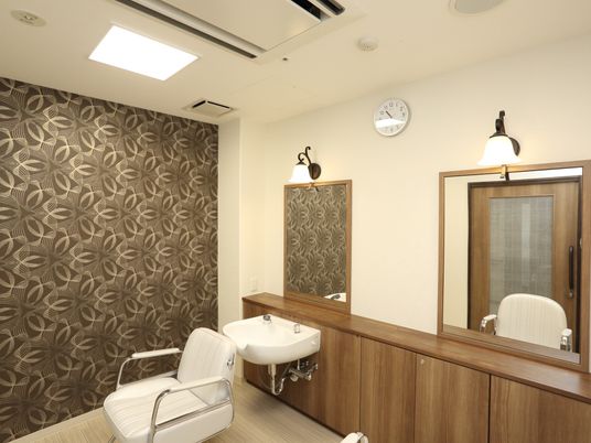 個性的なデザインの壁紙が施された部屋に、理美容専用チェアが据えられている。洗髪洗面化粧台が設置され、壁に四角い鏡が取りつけてある。