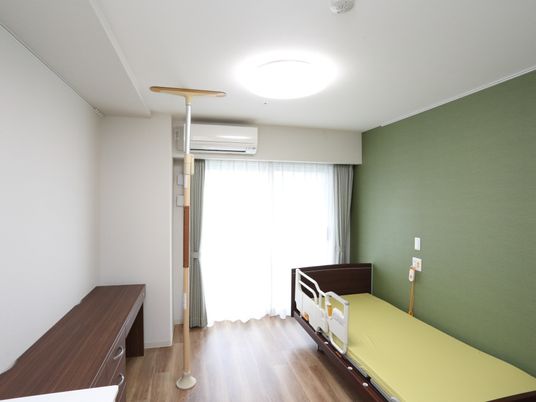 緑色と木目調が調和した部屋。ベッドと家具との間に、床と天井を利用して固定したポール状の手すりが置かれている。