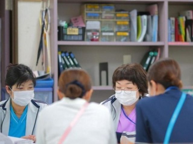 面談中のスタッフと利用者 多目的室   マスクを着けたスタッフが向かい合って話しており、落ち着いて相談しやすい雰囲気です。棚のある明るい空間で、打ち合わせや面談に使いやすそうです。