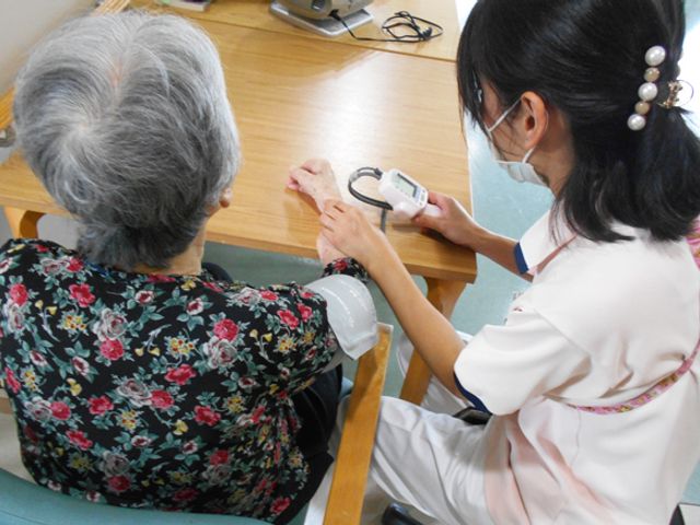 血圧測定の様子 機能訓練室 スタッフが測定器を使って、利用者の方の血圧のチェックを行っています。やさしい手つきで健康管理に努めています。