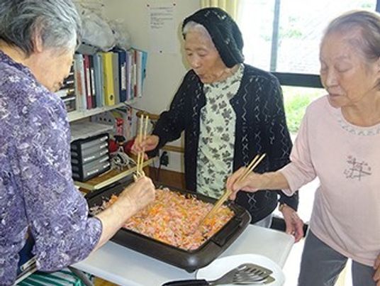 ホットプレートを囲んで料理をしている高齢女性たち。それぞれが菜箸を持っていて、そばにはフライ返しも置かれている。
