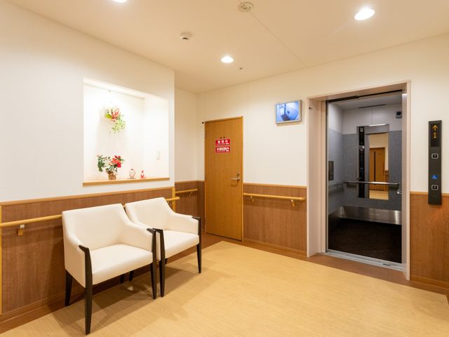 椅子が並ぶ明るい廊下 廊下 自然光が差し込む広々とした廊下で、エレベーターが近くにあります。壁には手すりも備えています。