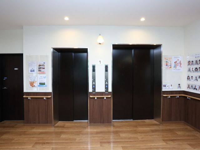 清潔なエレベーターホール エレベーター 床材に温もりを感じる木目調を採用し、情報掲示も充実しています。このホールで快適な移動をサポートします。