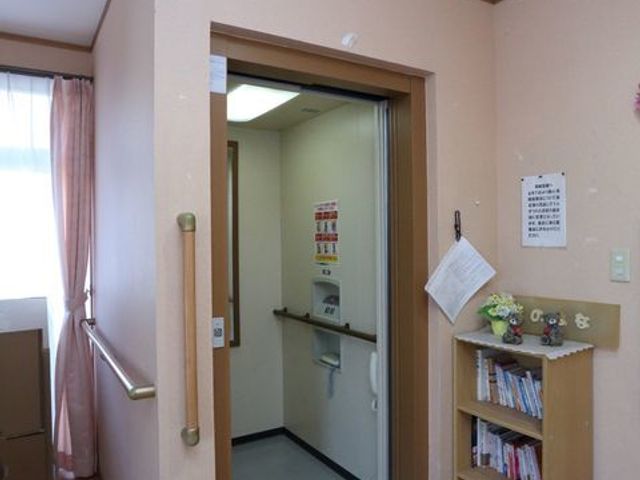 居室の入口と廊下 エレベーター ゆとりある入口は、利用者の安全と快適な移動を考慮して設計されています。壁には掲示物や飾り棚があり、暖かみのある雰囲気が感じられます。