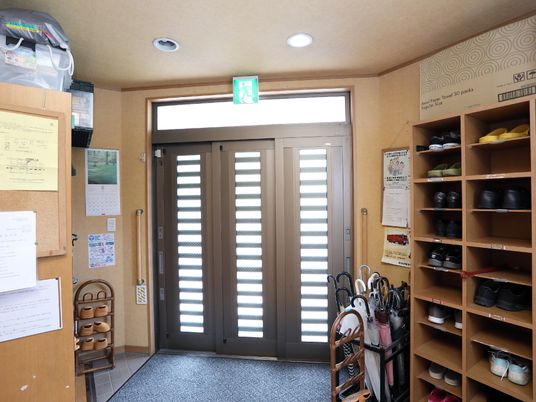 施設の写真 玄関は大きい靴箱が置かれている。手すりもあるので開閉や移動もしやすい構造。張り紙やカレンダーも貼られている。
