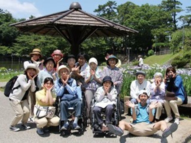 笑顔あふれる集合ポーズ 外出レク 外の清々しい景色の中で、スタッフと利用者様が仲良く記念撮影しています。温かみのある光景は、ここが働く場所としても心地よい環境であることを物語っています。
