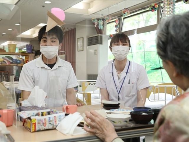 笑顔で接するスタッフと高齢者の様子 ダイニングルーム スタッフが笑顔で話しかける明るい空間です。多くの自然光が入り、居心地の良さが特徴です。